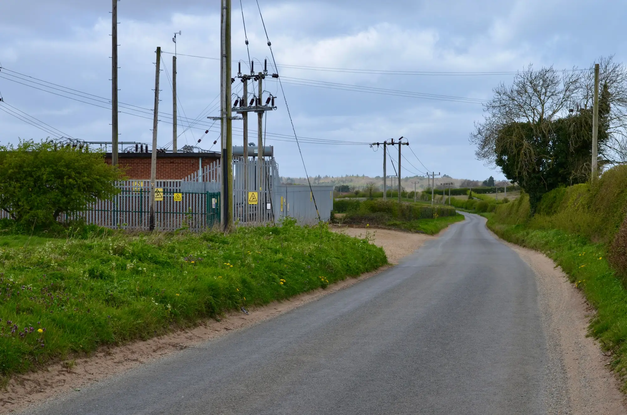 A paved country lane curving past a fenced electrical substation and a line of power poles